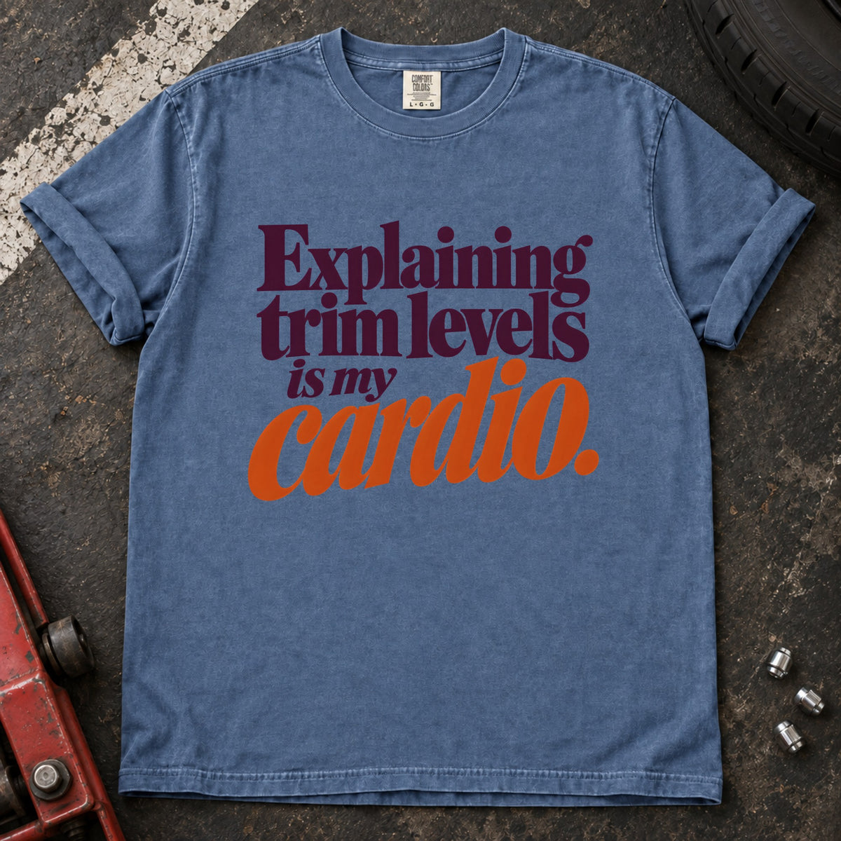 Blue Jean T-shirt with burgundy and orange lettering reading 'Explaining trim levels is my cardio'.