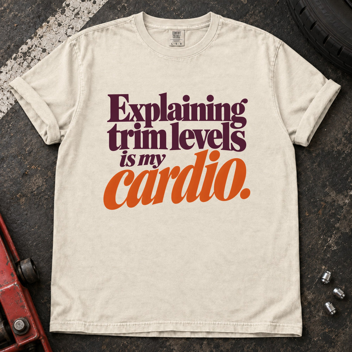 Ivory T-shirt with burgundy and orange lettering reading 'Explaining trim levels is my cardio'.